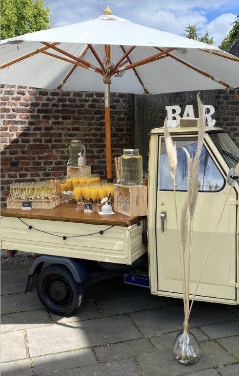 Ape-Bar mit Sonnenschirm vor einer alten Ziegelmauer, Theke mit Getränkespendern und Gläsern