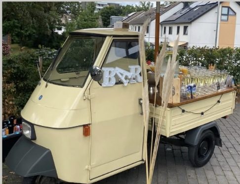 Frontansicht der cremefarbenen Piaggio Ape mit BAR-Buchstaben und Sektgläsern