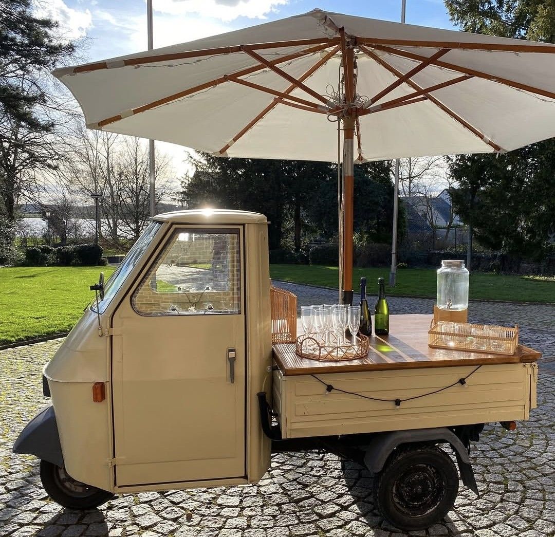 Livape – cremefarbene Piaggio Ape als mobile Bar mit Sonnenschirm, Sektgläsern und Holztheke