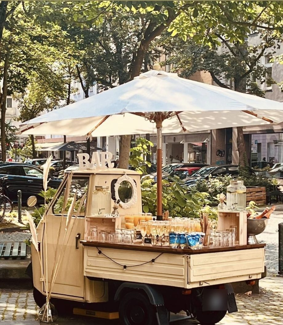 Piaggio Ape Bar von Livape mit Sonnenschirm und Sektgläsern auf einem Kopfsteinpflaster-Platz